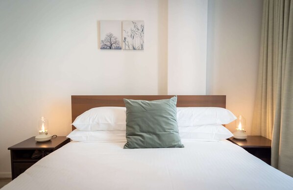 Una cama bien hecha con sábanas blancas y una única almohada verde, flanqueada por dos mesitas de noche con lámparas.