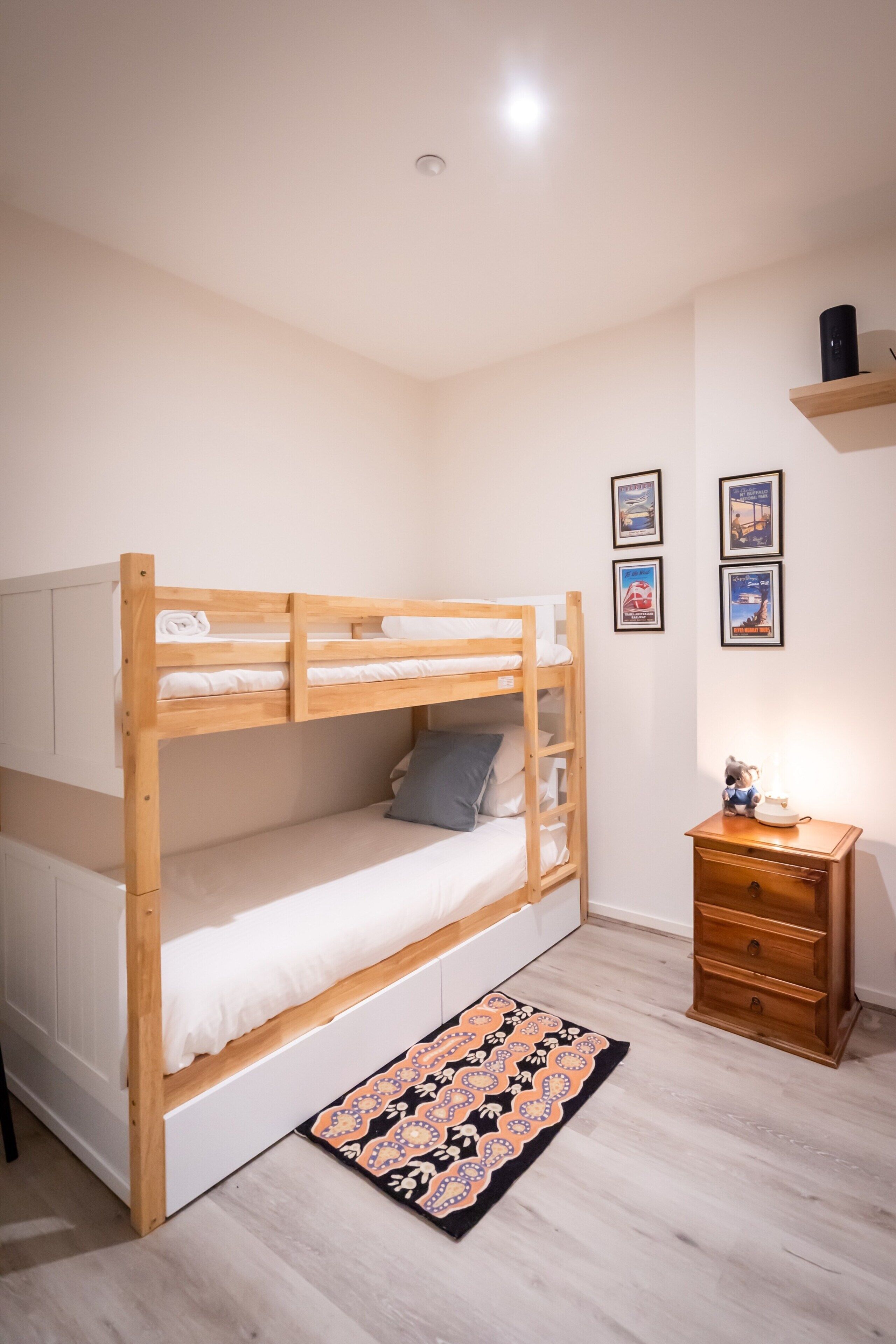 Ein Stockbett mit Holzrahmen und weißer Bettwäsche, ein kleiner Nachttisch mit Lampe sowie ein gemustertes Teppich auf hellem Holzboden.