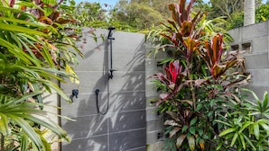 A modern outdoor shower area with a grey tiled wall, surrounded by lush greenery and plants. - Trendy 2-bed Home With Outdoor Entertainment (Sunshine Coast)