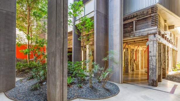 Un bâtiment moderne avec des piliers en béton, un espace jardin agrémenté de petits arbres et un escalier en bois menant à un niveau supérieur.