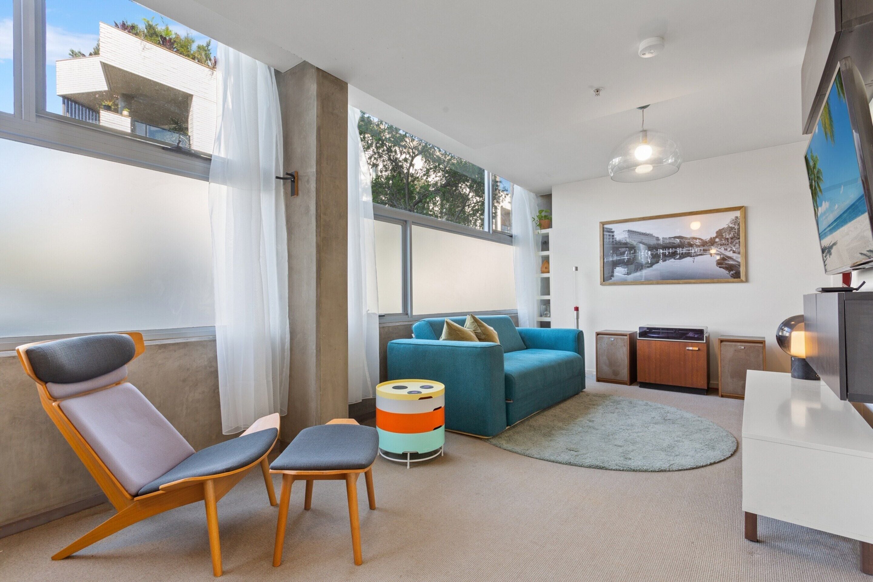 A modern living room with a blue sofa, a wooden chair, a colorful ottoman, and a flat-screen TV.
