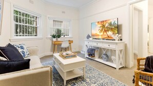 A living room with a white sofa, a coffee table, a TV, and a painting of palm trees. - Art Deco Apartment Meters Away From The Beach (Sydney)