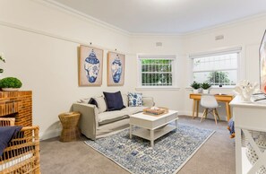 A living room with a sofa, coffee table, and a desk with a chair. There are two framed artworks on the wall, a window with a view of greenery, and a white ceiling. - Art Deco Apartment Meters Away From The Beach (Sydney)