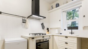 A compact kitchen with white cabinetry, a built-in oven, a gas stove, and a window with a view of palm trees. - Art Deco Apartment Meters Away From The Beach (Sydney)