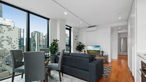 A modern living room with a grey sofa, a glass coffee table, and a dining area with chairs. There is a large window offering a city view. - Bright Apartment With City Views in Trendy Locale (Melbourne)