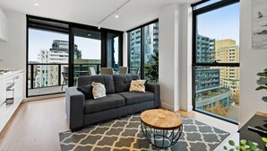 A modern living room with a grey sofa, a round coffee table, and large windows offering city views. - Bright Apartment With City Views in Trendy Locale (Melbourne)