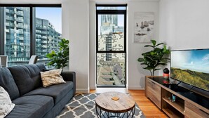 A modern living room with a grey sofa, a round coffee table, a TV on a wooden cabinet, and large windows offering a city view. - Bright Apartment With City Views in Trendy Locale (Melbourne)
