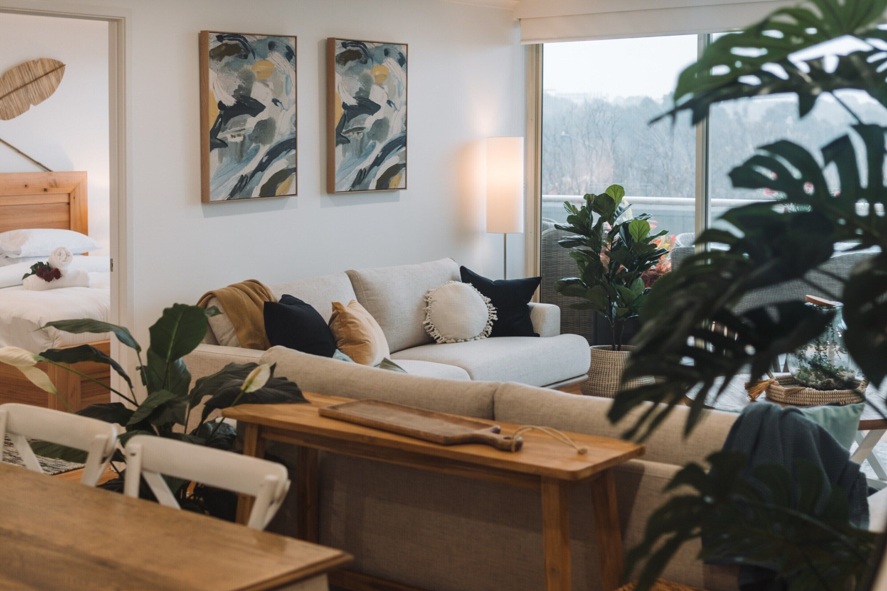 A modern living room with a sofa, a wooden table, and a dining area. There are two abstract paintings on the wall and a large window with a view of trees.