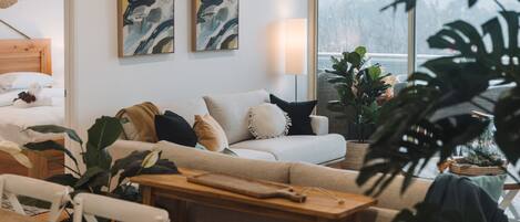 A modern living room with a sofa, a wooden table, and a dining area. There are two abstract paintings on the wall and a large window with a view of trees.