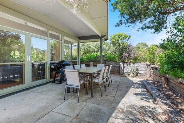 An outdoor patio with a table and chairs, a grill, and a covered area with large windows. - Spacious Coastal 4-bed Steps From the Sand (Melbourne)