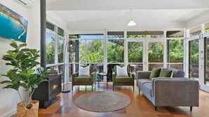 A spacious living room with a fireplace, green armchairs, a grey sofa, and large windows offering a view of greenery. - Spacious Coastal 4-bed Steps From the Sand (Melbourne)