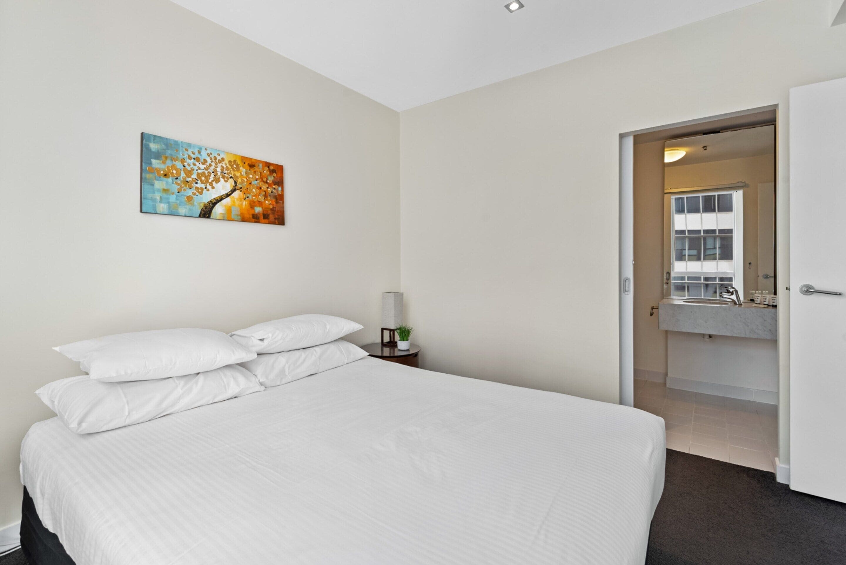 A hotel room with a large bed, a nightstand with a lamp and a small plant, a painting on the wall, and a bathroom visible through an open door.