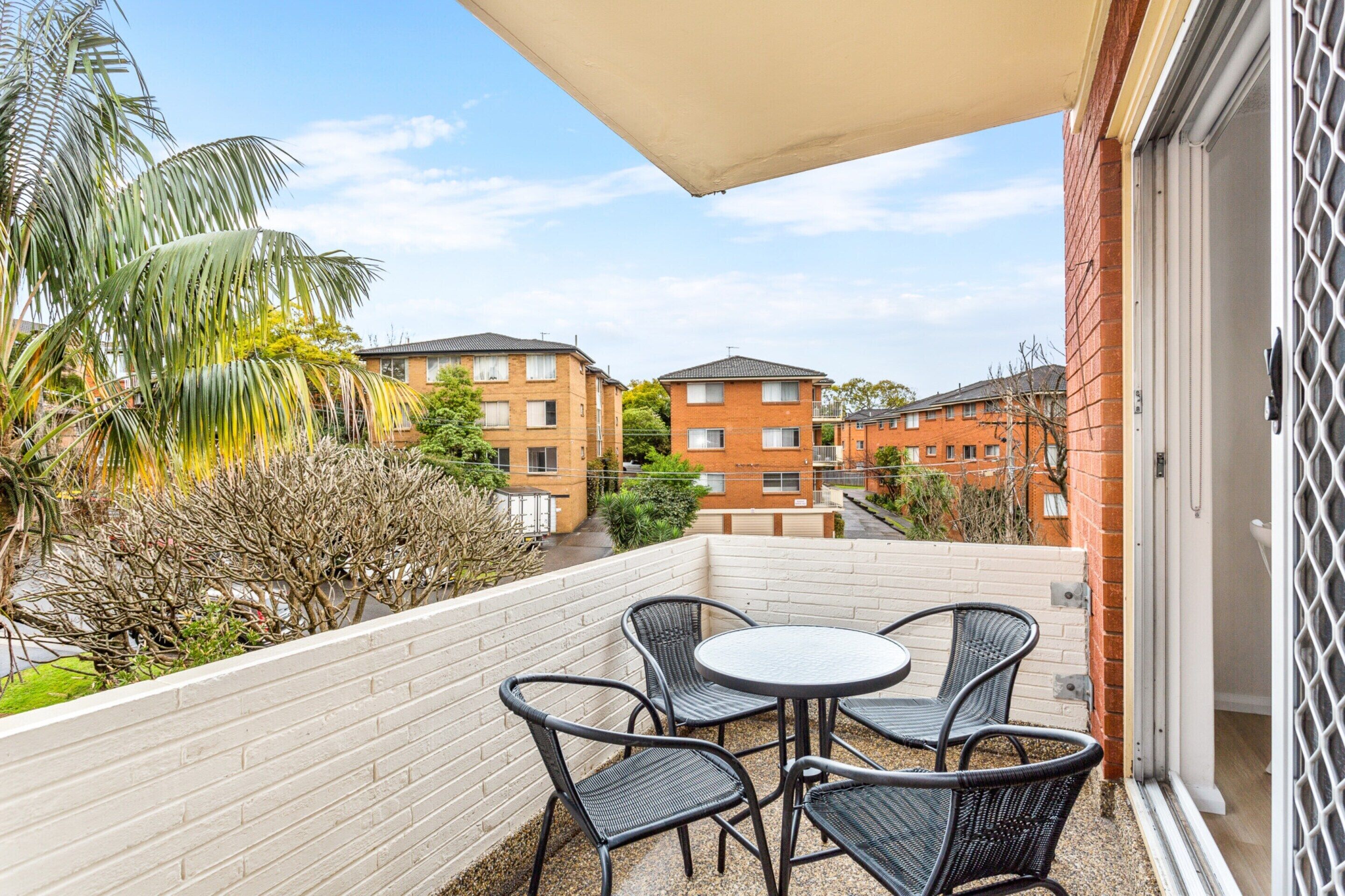 A balcony with a table and chairs, a white fence, and buildings in the background.