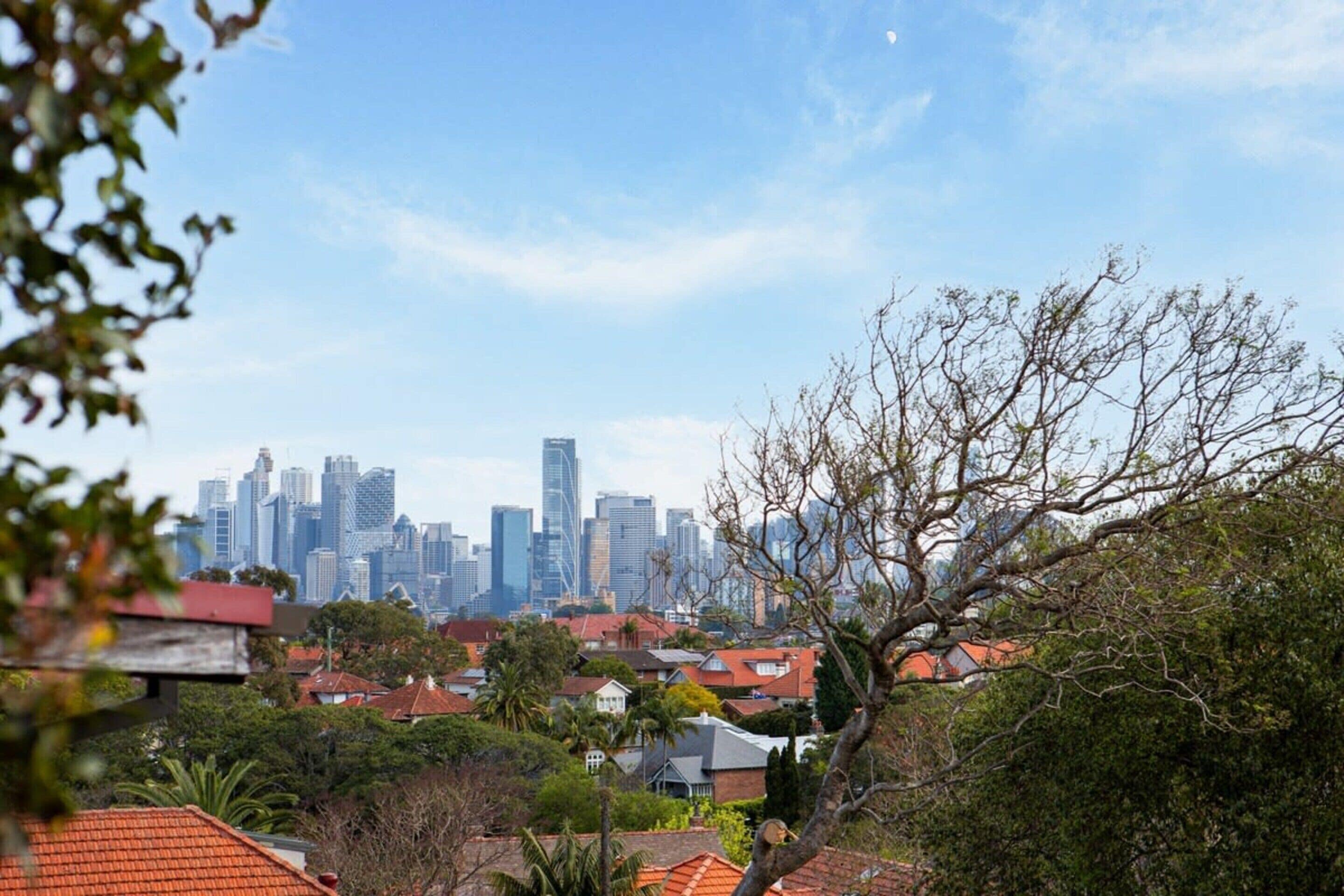 Une vue sur les toits d’une ville, avec des immeubles modernes de grande hauteur, entourés de maisons résidentielles aux toits de tuiles rouges et d’une végétation luxuriante.