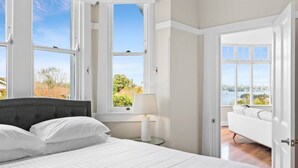 A bedroom with a large bed, a nightstand with a lamp, and a view of a cityscape through large windows. - Huge Harbour View Apartment In Historic Home (Sydney)