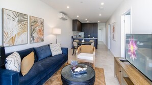 Una sala de estar moderna con un sofá azul oscuro, una mesa de centro de vidrio y un televisor de pantalla plana. Hay una zona de comedor con una mesa y sillas, y una cocina al fondo.