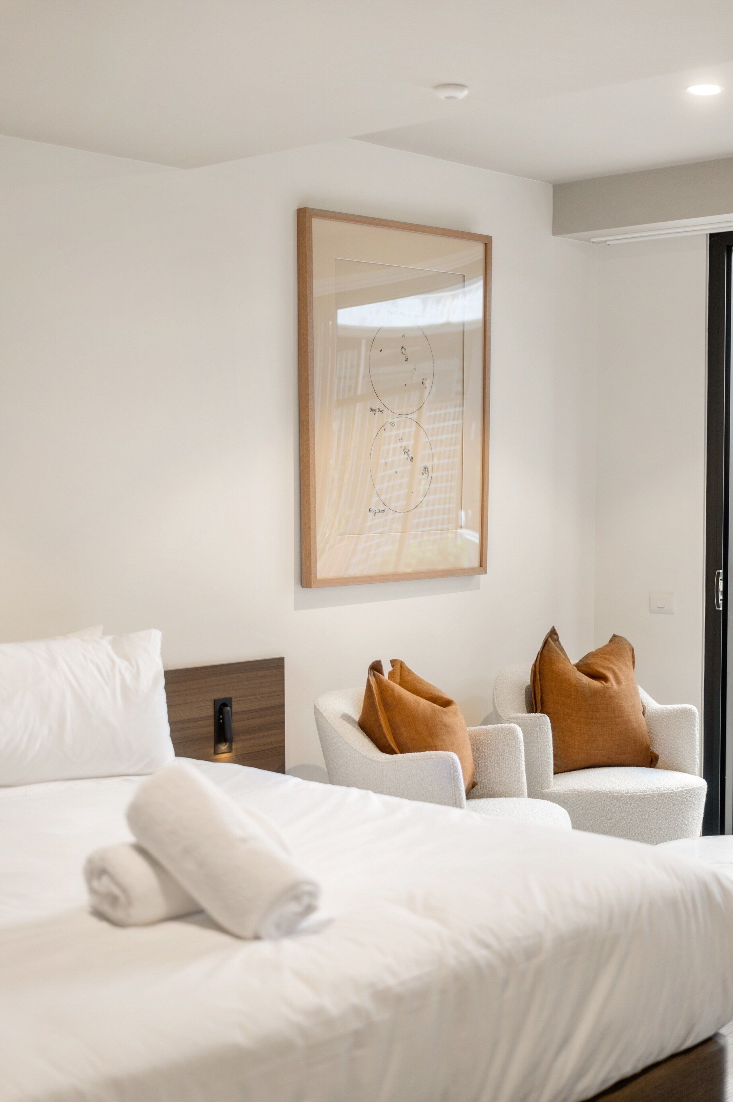 A modern hotel room with a neatly made bed, a seating area with two chairs, and a framed artwork on the wall.