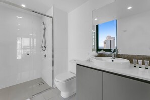 Et moderne badeværelse med bruseniche, hvidt toilet, stor vask med toiletartikler og et spejl der reflekterer et bybillede.