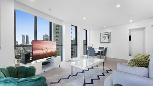 A modern living room with a large TV, green and white furniture, a glass coffee table, and a city view through large windows. - Wonderful 2-bed With Pool, Gym & Scenic Views (Gold Coast)