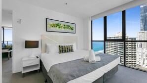 A modern hotel room with a large bed, a nightstand, a lamp, and a view of a cityscape through large windows. - Wonderful 2-bed With Pool, Gym & Scenic Views (Gold Coast)