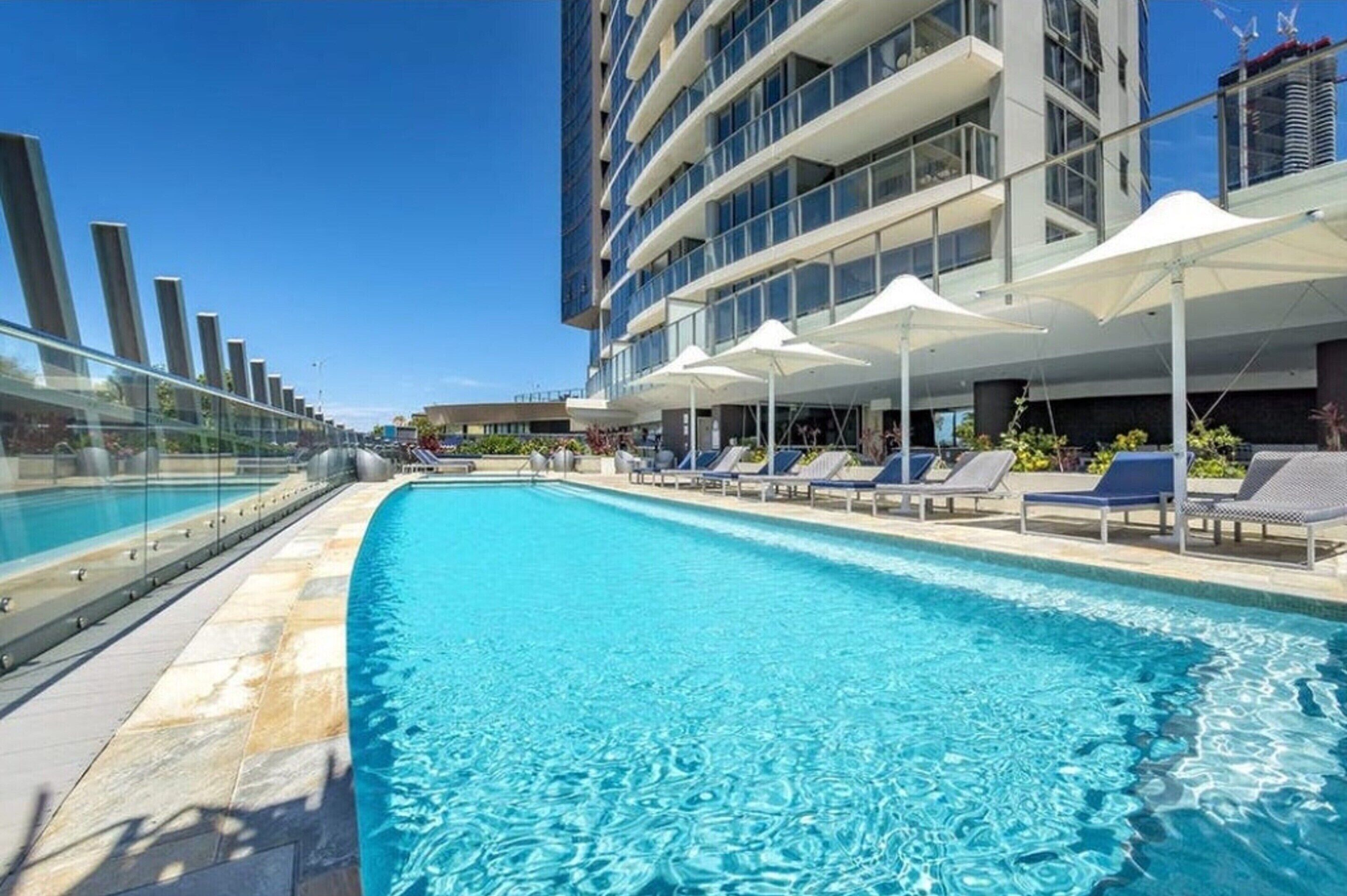 Una piscina con lettini e ombrelloni, circondata da un edificio moderno con balconi.