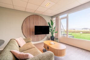Un salón moderno con un televisor grande montado en una pared de paneles de madera, un sofá cómodo, una mesa de centro de madera y vistas al exterior a través de amplios ventanales.