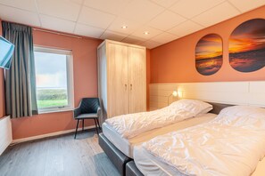 Una habitación de hotel con cama, una silla, armario, televisor, ventana con cortinas y un cuadro de atardecer en la pared.