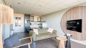 A modern living room with a sofa, chairs, a round wooden table, and a TV mounted on a wooden panel wall. - Sea View Apartment (De Koog)