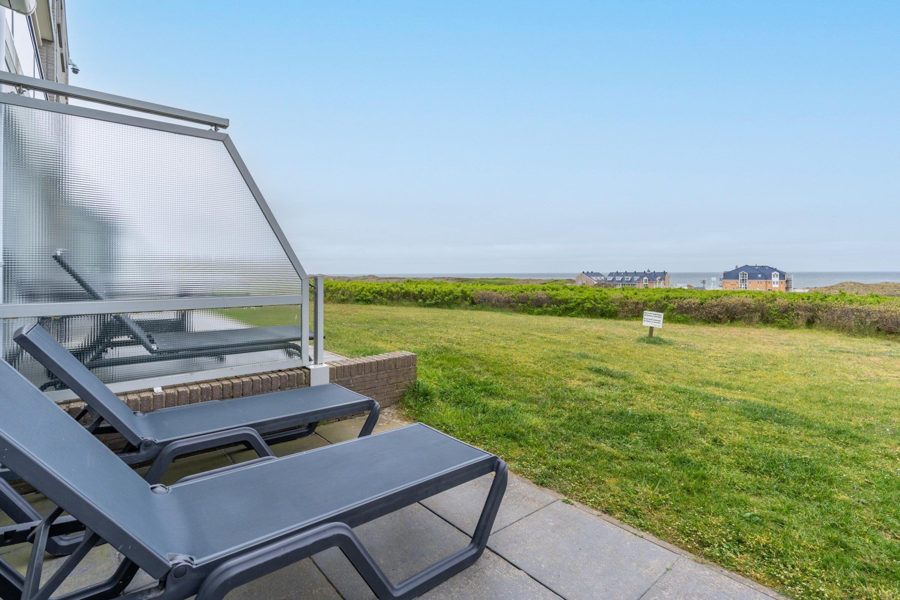 A patio area with a view of the sea, a grassy lawn, and a building in the background.