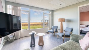 A modern hotel room with a large window offering a view of the countryside, a flat-screen TV, a comfortable seating area with a sofa and chairs, and a small round table. - Sea View Apartment (De Koog)