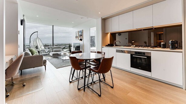 A modern kitchen with white cabinets, a dining area with a round table and chairs, and a city view through large windows. - 2-bed With Harbour Views & Outstanding Amenities (Melbourne)