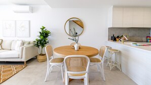A modern living room with a round wooden dining table, white wicker chairs, a white sofa, and a large mirror on the wall. - Lavish Coastal 2-bed With Stunning Ocean Views (Batemans Bay)