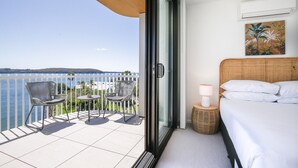 A balcony with a view of the sea, a bed with white bedding, wicker chairs, and a small table. - Lavish Coastal 2-bed With Stunning Ocean Views (Batemans Bay)