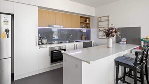 A modern kitchen with white cabinetry, a built-in oven, a stainless steel refrigerator, and a bar area with stools. - Spaciously Bright 1-bed Apartment (Brisbane)