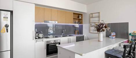 A modern kitchen with white cabinetry, a built-in oven, a stainless steel refrigerator, and a bar area with stools.