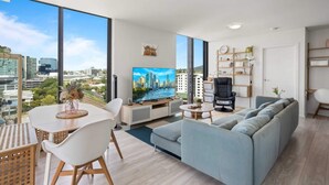 A modern living room with a large TV, a sofa, a dining table with chairs, and a city view through large windows. - Spaciously Bright 1-bed Apartment (Brisbane)