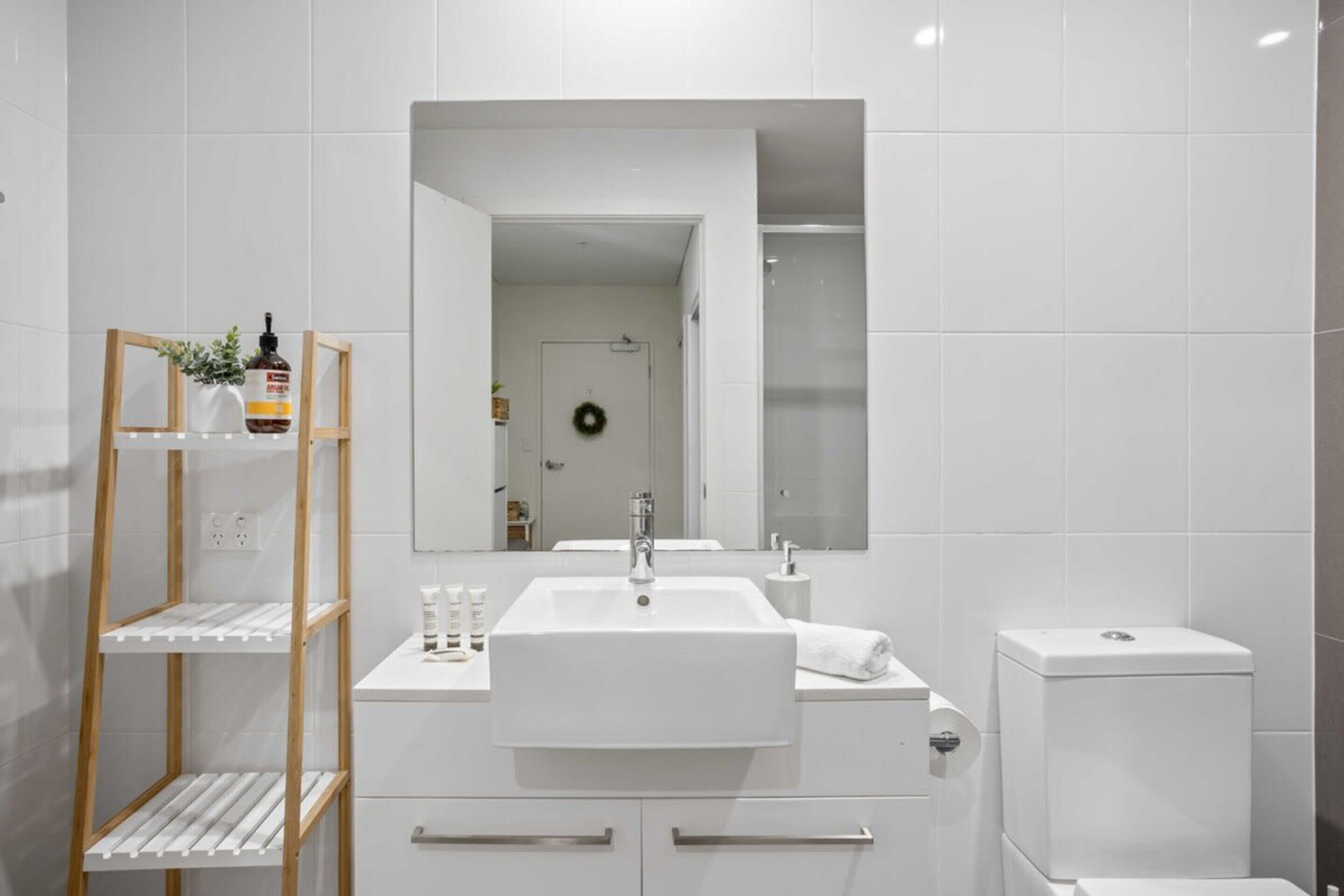 A modern bathroom with a white sink, mirror, and toilet. There is a wooden shelf with toiletries and a small plant.