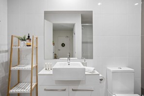 Une salle de bain moderne avec un lavabo blanc, un miroir et des toilettes. On y trouve une étagère en bois avec des articles de toilette et une petite plante.