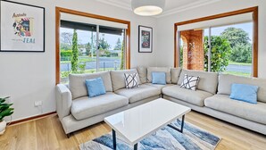 A modern living room with a sectional sofa, a coffee table, and large windows offering a view of the outdoors. - 4-bed Beach House With Garden & Alfresco Dining (Tomakin)