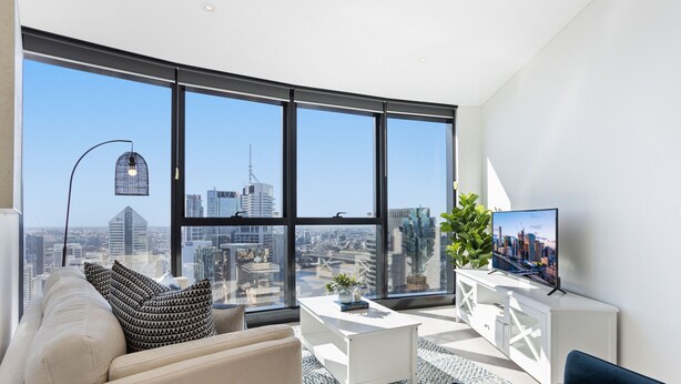 Un salon moderne avec vue sur la ville, un canapé blanc, une table basse et un téléviseur posé sur un meuble blanc.