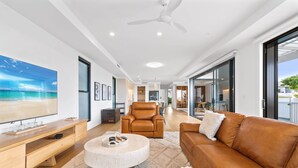 A modern living room with a large flat-screen TV, leather sofas, a round coffee table, and a ceiling fan. - Deluxe 4-bed Home With Pool & Fire Pit (Brisbane)