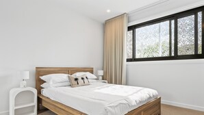 A bedroom with a wooden bed, white bedding, a nightstand with a lamp, a window with a view of greenery, and a white wall. - Deluxe 4-bed Home With Pool & Fire Pit (Brisbane)