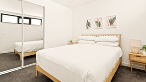 A bedroom with a large mirror, a bed with white bedding, a nightstand with a lamp, and three framed botanical prints on the wall.