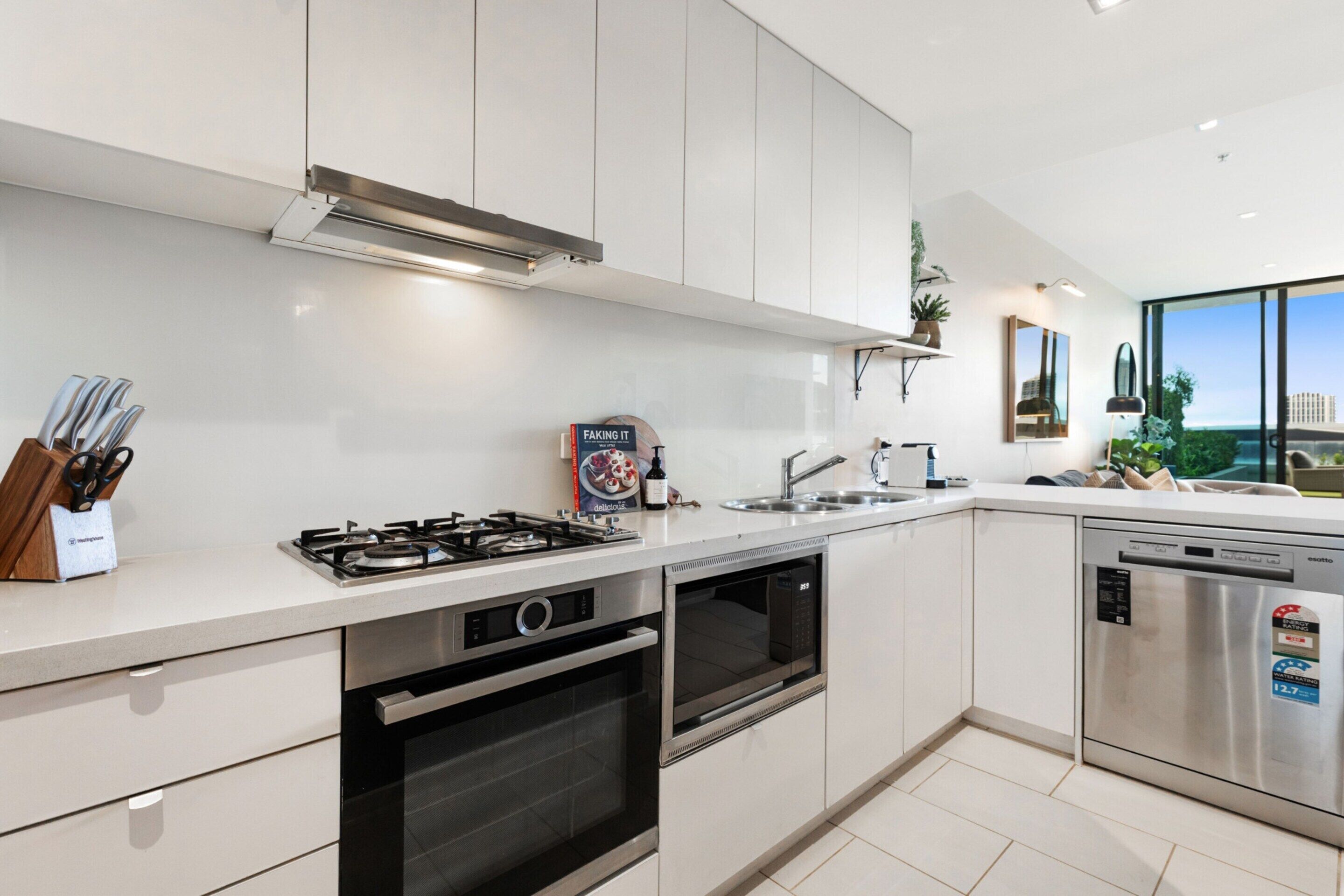 Une cuisine moderne avec des meubles blancs, une cuisinière à gaz, un four, un lave-vaisselle et une grande fenêtre donnant sur la ville.