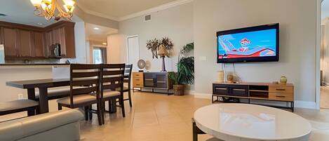 A spacious living room with a large TV, a dining area with wooden furniture, and a kitchen with wooden cabinets.