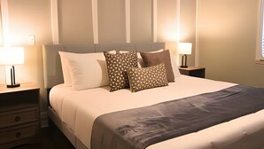 Un lit bien fait, recouvert d’une couverture grise, deux tables de chevet avec des lampes et une fenêtre avec des stores.