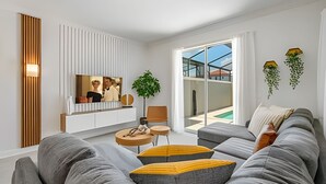 Un salon moderne avec un canapé gris, des poufs jaunes, un téléviseur fixé à un mur blanc et une grande fenêtre donnant sur une piscine et des immeubles.