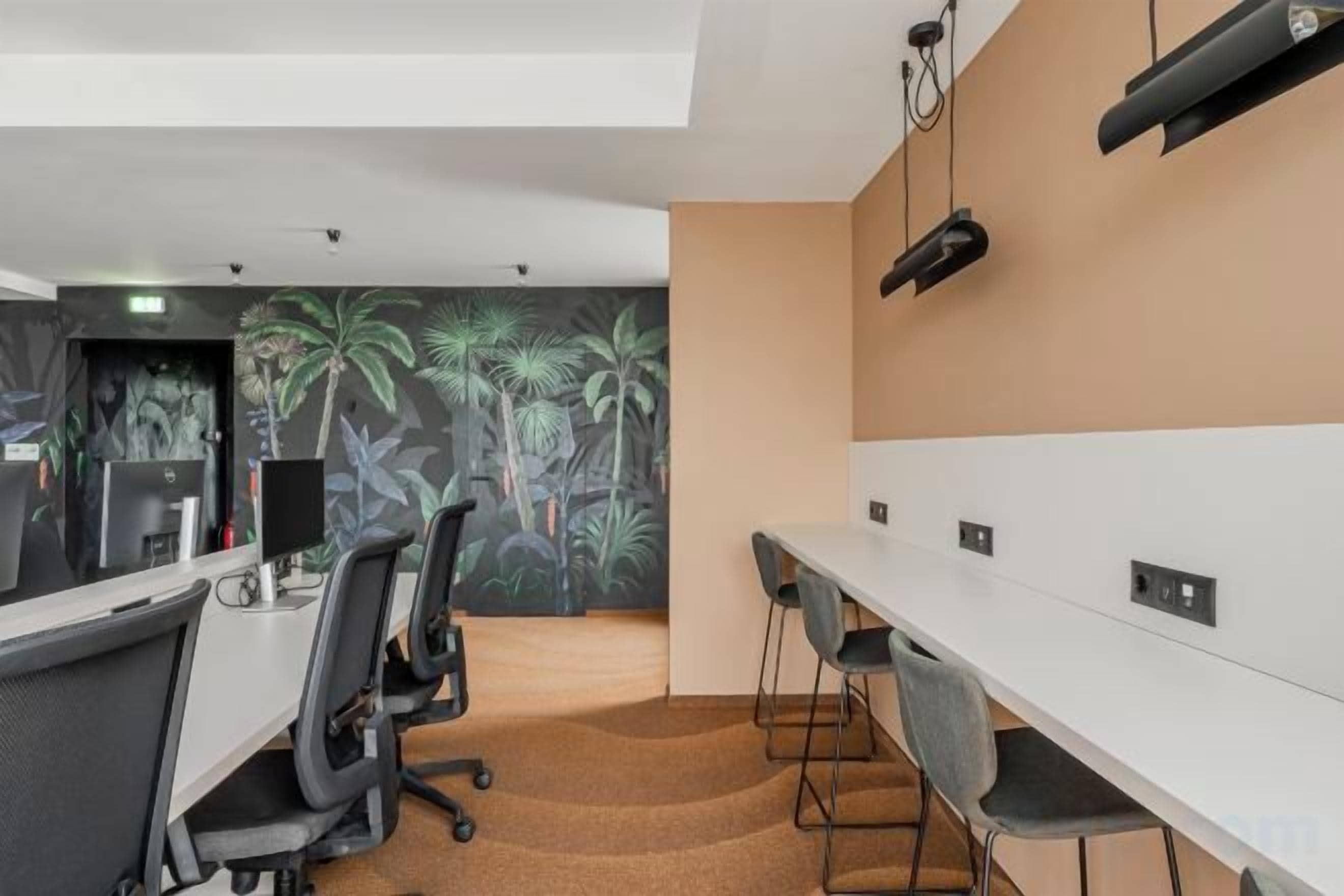 Un espacio de oficina moderno con mesas largas, sillas y un mural con follaje tropical en la pared.