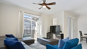 En moderne stue med sofa, skammel og tv på et stativ. Der er ventilator i loftet, en skydedør af glas og et spiseområde med en stol.