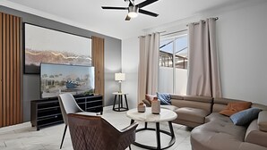 A modern living room with a flat-screen TV, a ceiling fan, a sofa, and a coffee table. - Disney 4BR w/ Pool + Themed Rooms in Storey Lake (Kissimmee)